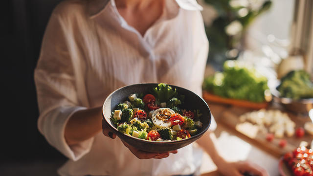 Woman in t-shirt and jeans standing and holding vegan superbowl or Buddha bowl with hummus, vegetable, salad, beans, couscous and avocado and smoothie in hands, square crop 